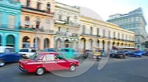 A small iconic red classic old car fast moving speeding fast along Paseo de Marti with colourful colonial buildings, Havana, Cuba