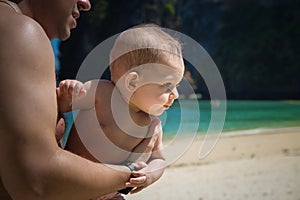 A small infant baby in the arms of dad, very much interested, turned away. Reaches for mom. Toddler on sunny tropical beach