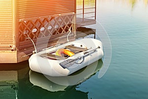 Small inflatable row boat with paddles moored near wooden gazebo on pier. Fishing boat tied at lake or river shore on early