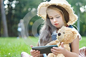 Small kid girl looking in her mobile phone together with her favorite teddy bear toy outdoors in summer park