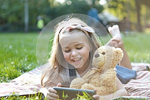 Small kid girl looking in her mobile phone together with her favorite teddy bear toy outdoors in summer park