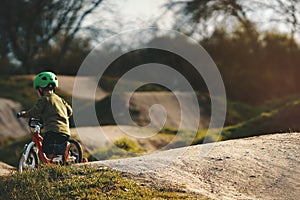 small kid with green helmet rides a balance bike on a dirt jump track in bright sunlight