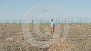 Small lonely child goes state border. Resentful boy from a refugee settlement holds a toy in his hand, go away and turns around in