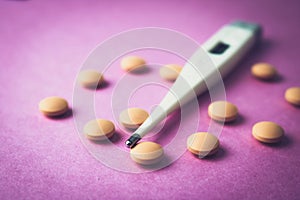 Small medical pharmacetic round pills, vitamins, drugs and electronic digital thermometer on a pink purple background.