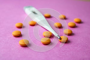 Small medical pharmacetic round pills, vitamins, drugs and electronic digital thermometer on a pink purple background.