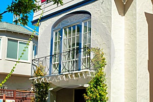 Small metal decorative balcony with white building facade on stucco house with vines and front yard trees and foliage