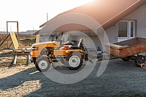 Small mini red modern orange new tractor with trailer standing near hangar building at farm countryside during sunset or