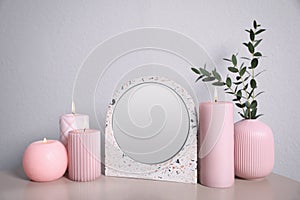 Small mirror, burning candles and vase on table near light wall