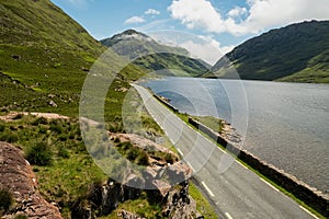 Small narrow road in a mountains by a lake with blue water with beautiful nature scenery. Warm sunny day, cloudy sky. Connemara