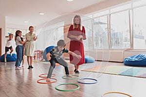 Small nursery school children with female teacher on floor indoors in classroom, doing exercise. Jumping over hula hoop