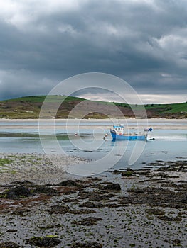A small old fishing boat is anchored, shallow water at low tide on a cloudy day. Hills