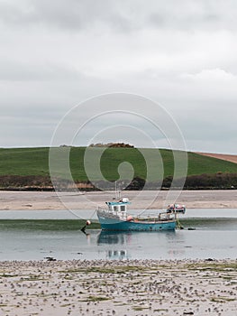A small old fishing boat is anchored, shallow water at low tide on a cloudy day. Hills under a cloudy sky. Silt and seaweed.