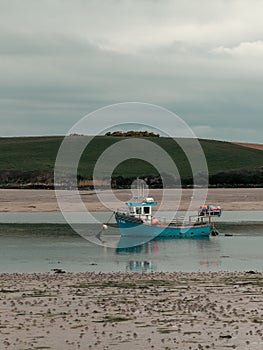 A small old fishing boat is anchored, shallow water at low tide on a cloudy day. Hills under a cloudy sky. Silt and seaweed