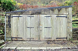 Small old stone storage or garage building with decaying wooden doors and rusted hinges with damp walls and roof