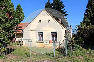 Small old suburban family house with two windows and decorative attic ventilation openings surrounded with rusted metal fence