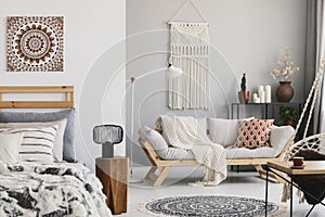 Small open space flat interior with beige sofa with cushion, macrame on the wall, rack with candles and plants and bed with