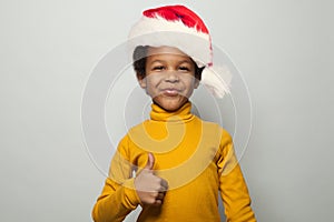 Small optimistic black child boy in Santa hat smiling and showing thumb up on white background