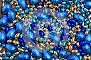 Small pale and dark blue, silver and gold foil wrapped chocolate easter eggs, with large blue eggs against a pale background.