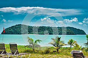 Small palm trees and deck chairs looking at a tropical island in the azure sea under the blue sky on the shores of the sandy