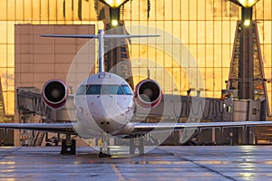 A small plane stands on the apron platform on the background of a glass terminal building with sunset light reflected in the sky