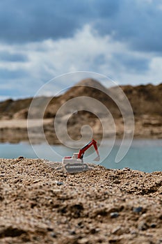 Small plastic toy excavator with bucket working on sand extraction at quarry. Pond in background. Children`s toy model of tractor