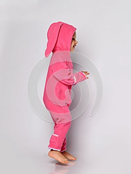 Small positive barefoot cute blonde baby girl in pink warm comfortable jumpsuit with hood has fun jumping. Side view