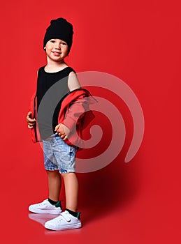 Small positive boy in denim shorts, red jacket, cap white sneakers standing, smiling and looking aside over red wall background