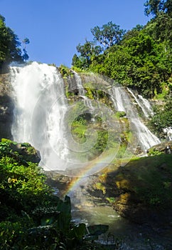 A small rainbow below a waterfall surrounded by green plants