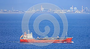 Small red cargo ship sails past chimneys at industrial refinery on coast