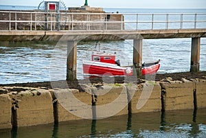 A small red fishing boat passes a pier on a calm day