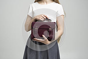 Small red leather bag in a woman& x27;s hand on a white background. Shoulder handbag. Woman in a white shirt and grey skirt