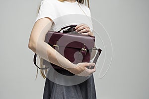 Small red leather bag in a woman& x27;s hand on a white background. Shoulder handbag. Woman in a white shirt and grey skirt