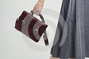 Small red leather bag in a woman's hand on a white background. Shoulder handbag. Woman in a white shirt and grey skirt
