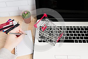 Small red shopping cart or trolley on laptop keyboard and woman hand writing in notes. Technology business online shopping concept