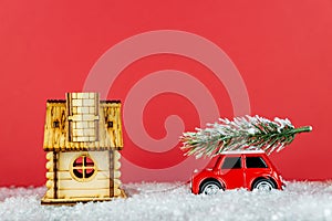 Small red toy car carrying spruse tree on a snowe road next to wooden hous. Festive christmas greeting card