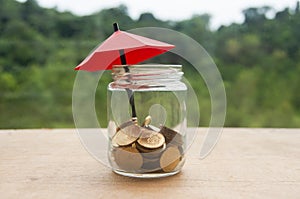 Small red umbrella on pile of gold coins inside a jar on wooden table. Financial safety and investment concept.