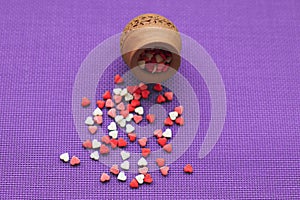 Small red, white and pink hearts scattered from a wooden vase on a violet background.