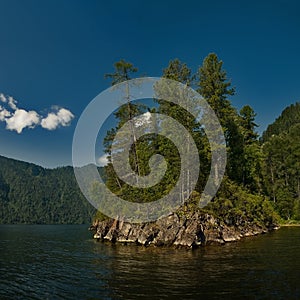 Small rocky islet overgrown with trees near the shore of the lake against the blue sky