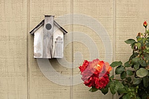 Small rustic handmade birdhouse decorated with a wood lighthouse it hanging next to beautiful orange, yellow, red rose bush