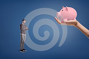 A small sad bearded man stands near a large female hand holding a piggy bank.