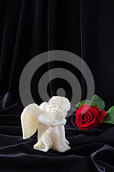 A small sculpture of a baby angel with wings and a red rose on a black background. The concept of funerals, condolences, and