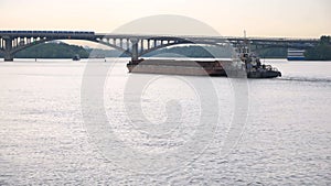 A small ship tows a barge with a cargo on the river in the direction of the bridge.