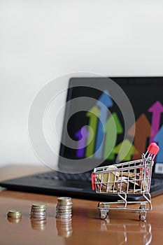 Small shopping cart full of coins on the table with stacks of coins and a laptop on it