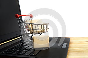 A small shopping cart with miniature shipping boxes stands on a laptop against a white background. Online shopping concept