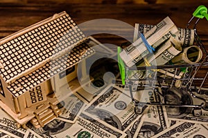 Small shopping cart with rolled up one hundred dollar banknotes and plywood model of the house on a wooden background. Loan, real