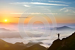 Small silhouette of hiker man enjoying beautiful sunrise in morning mountains. Traveler with raised hands standing on mountain