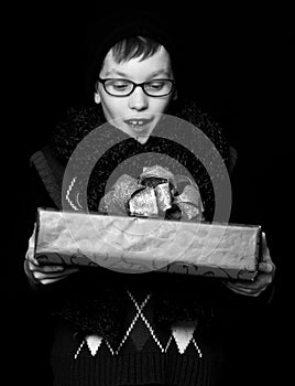 Small smiling boy or cute nerd kid in glasses, hat and fashionable knitted scarf on black background holds red present