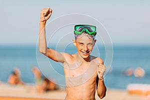 Small smiling child boy in swimming glasses, having fun and celebrating victory on the sea sand of the beach. Summer holiday