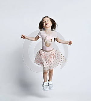 Small smiling cute girl in casual summer dress and sneakers jumping over white wall background