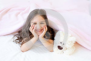 A small smiling girl 5-6 years old is lying in bed with a Teddy bear, hands under her cheeks